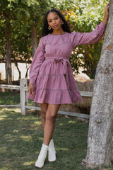 woman wearing a pink tiered dress with long sleeves and a tie waist, standing outdoors by a tree