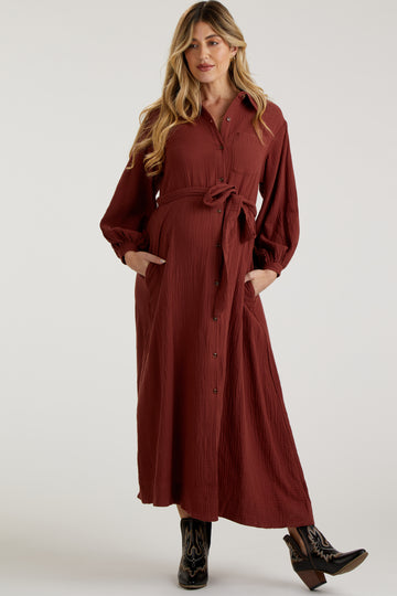 woman wearing a rust long sleeve maternity dress with a tie waist and buttons, standing with her hands in pockets and a visible baby bump