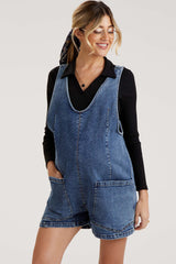 woman wearing a blue denim maternity romper with large front pockets and a black long sleeve top underneath