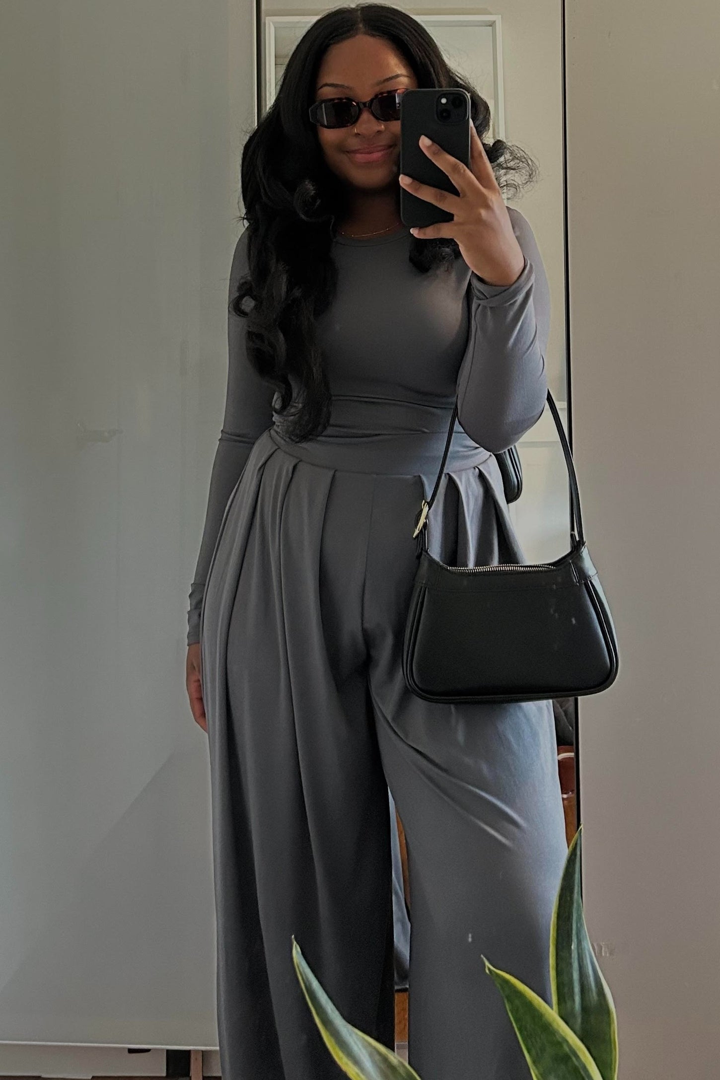 woman wearing a grey long sleeve jumpsuit with wide leg pants and holding a black handbag, posing in a mirror