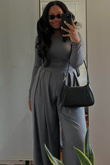 woman wearing a grey long sleeve jumpsuit with wide leg pants and holding a black handbag, posing in a mirror