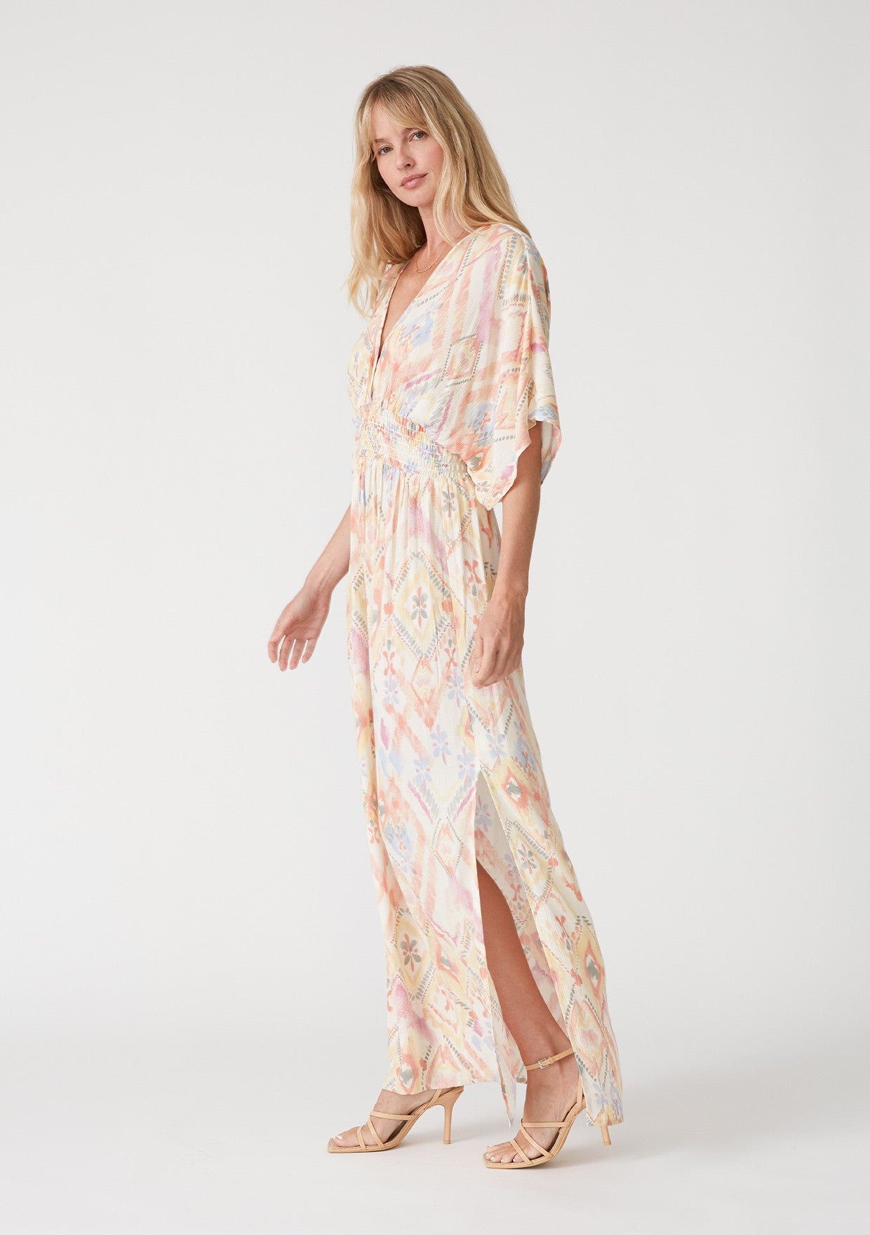 woman wearing a pastel-colored maxi dress with a geometric pattern and side slits, standing in profile with one hand on her hip