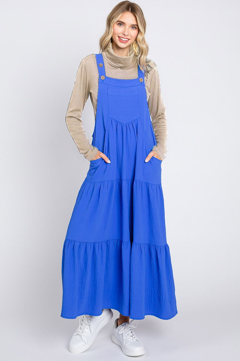 woman wearing a long blue tiered maxi dress with pockets and adjustable straps, styled with a beige turtleneck and white sneakers