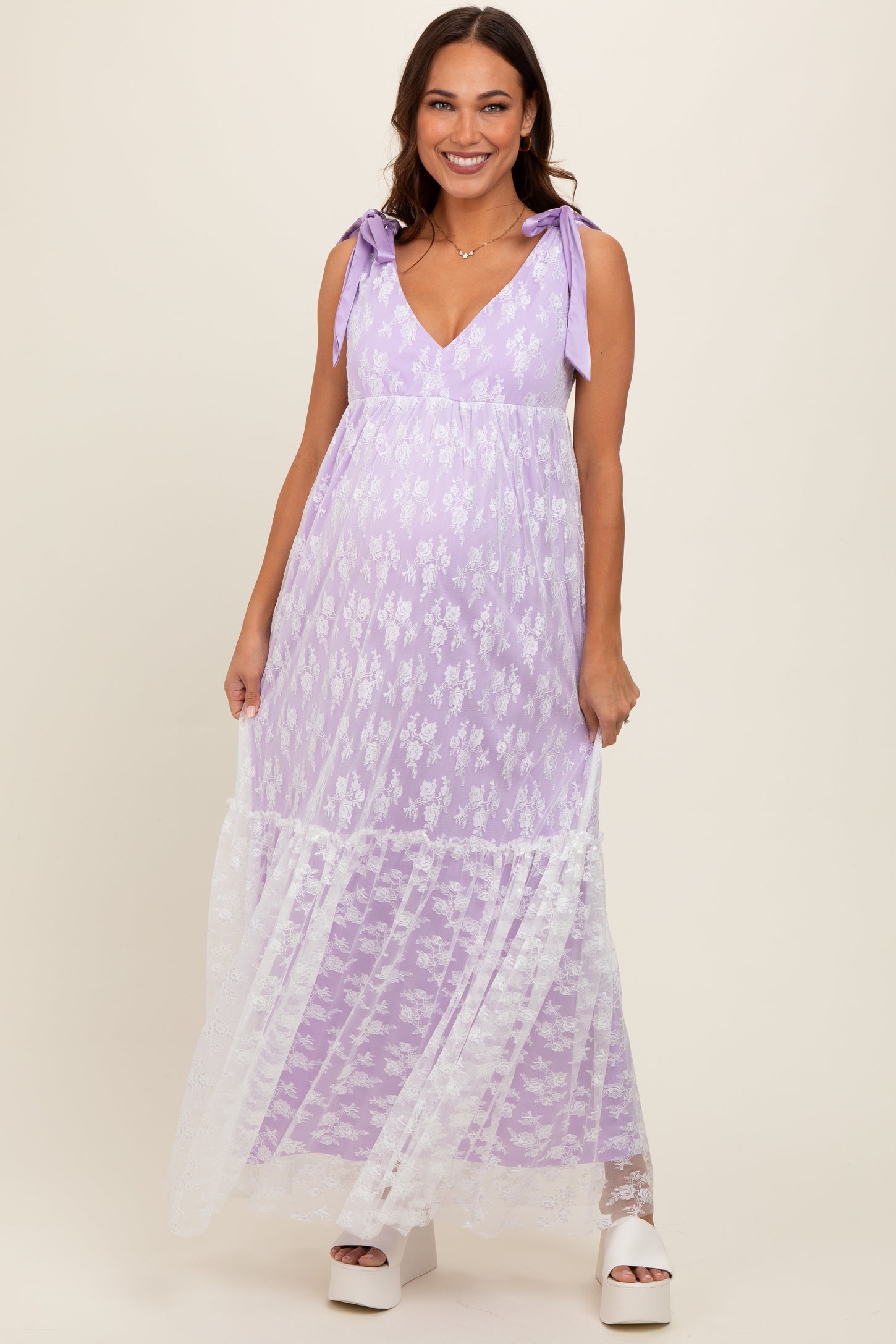 woman wearing a lavender maternity maxi dress with floral details and lace accents, holding the dress while smiling