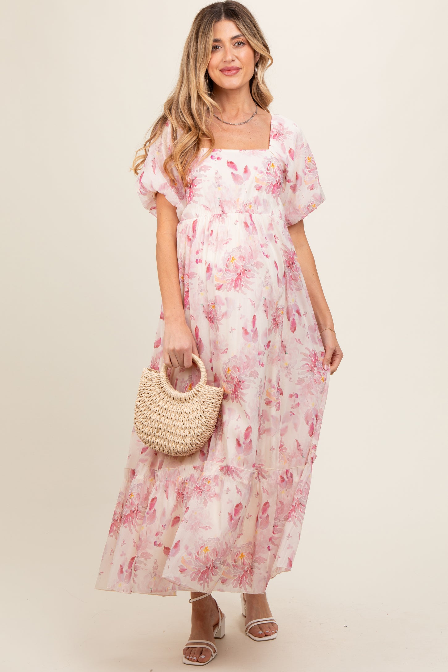 woman wearing a pink floral maternity maxi dress with puff sleeves and holding a woven bag