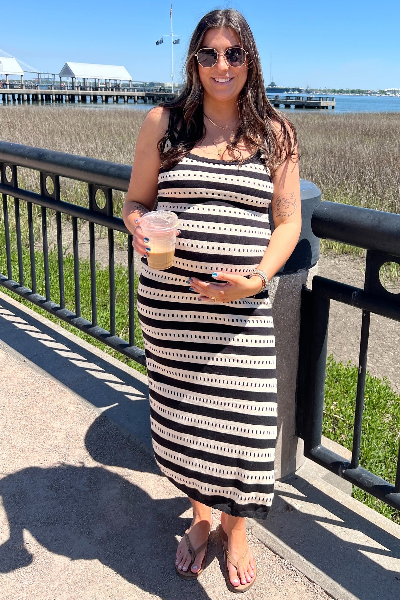 woman wearing a black and white striped maternity maxi dress while holding a drink by the waterfront