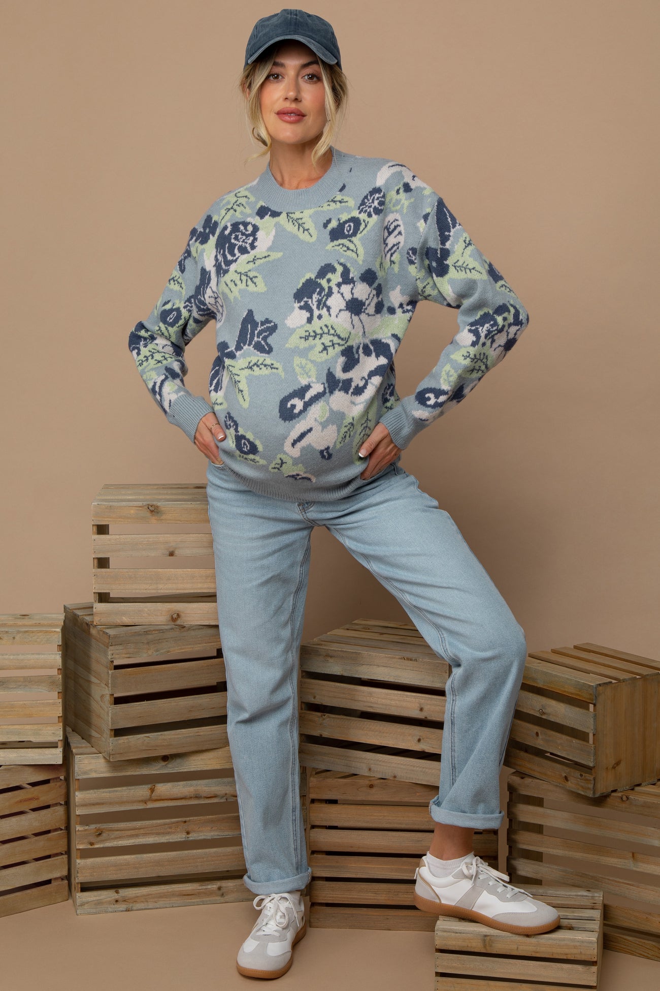 woman wearing a floral patterned light blue maternity sweater and light blue jeans, posing with a baby bump visible