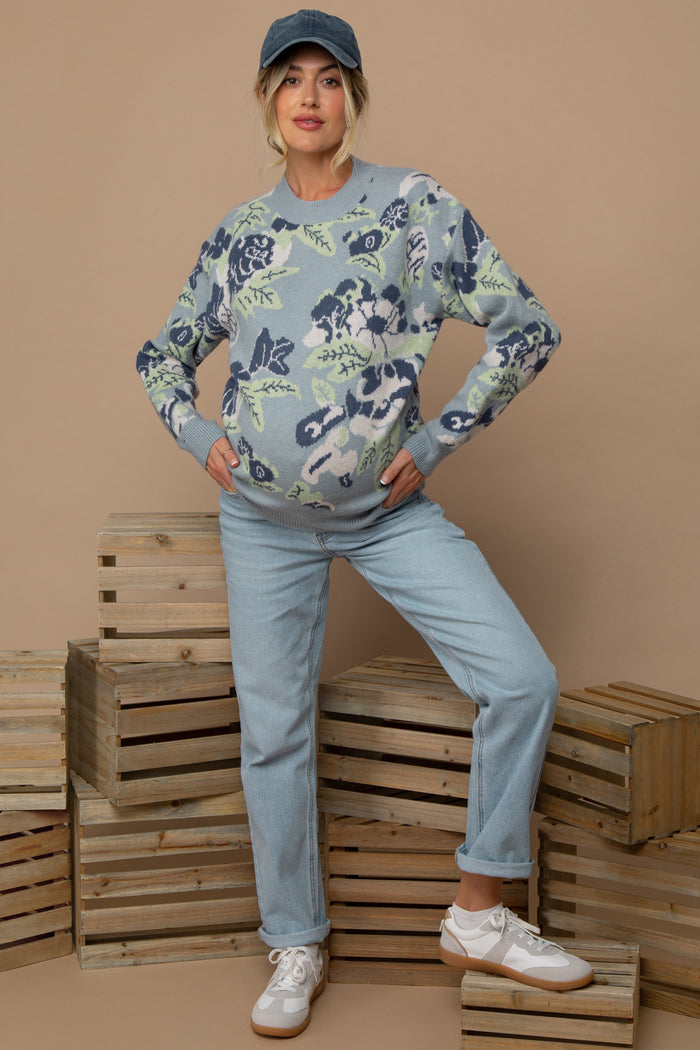 woman wearing a floral patterned light blue maternity sweater and light blue jeans, posing with a baby bump visible