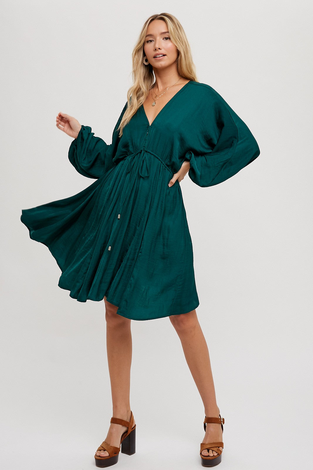 woman wearing a teal v-neck dress with long sleeves and a tie waist, posing with a playful spin