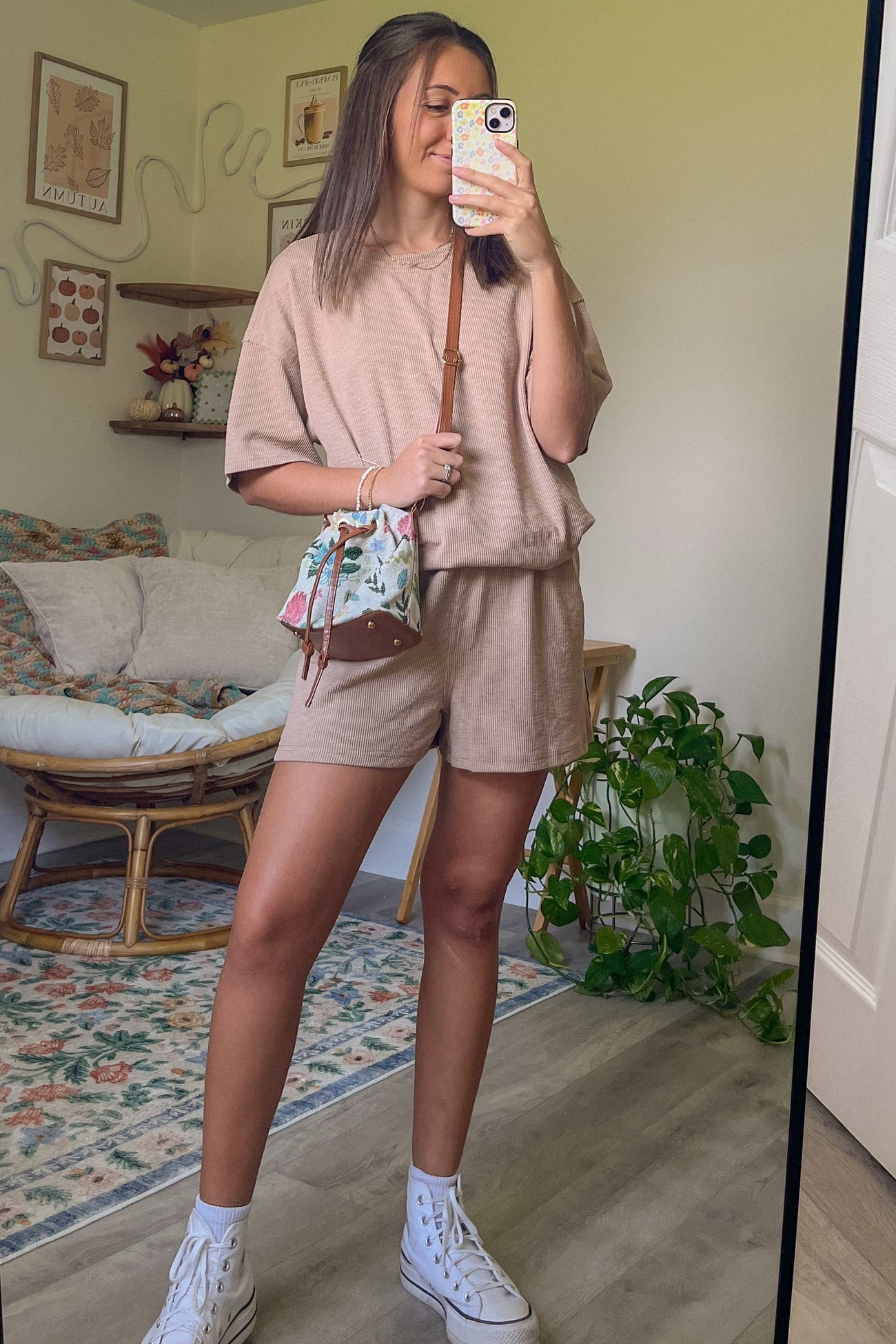 woman wearing a beige oversized short sleeve top and matching shorts, holding a floral handbag, standing in front of a mirror