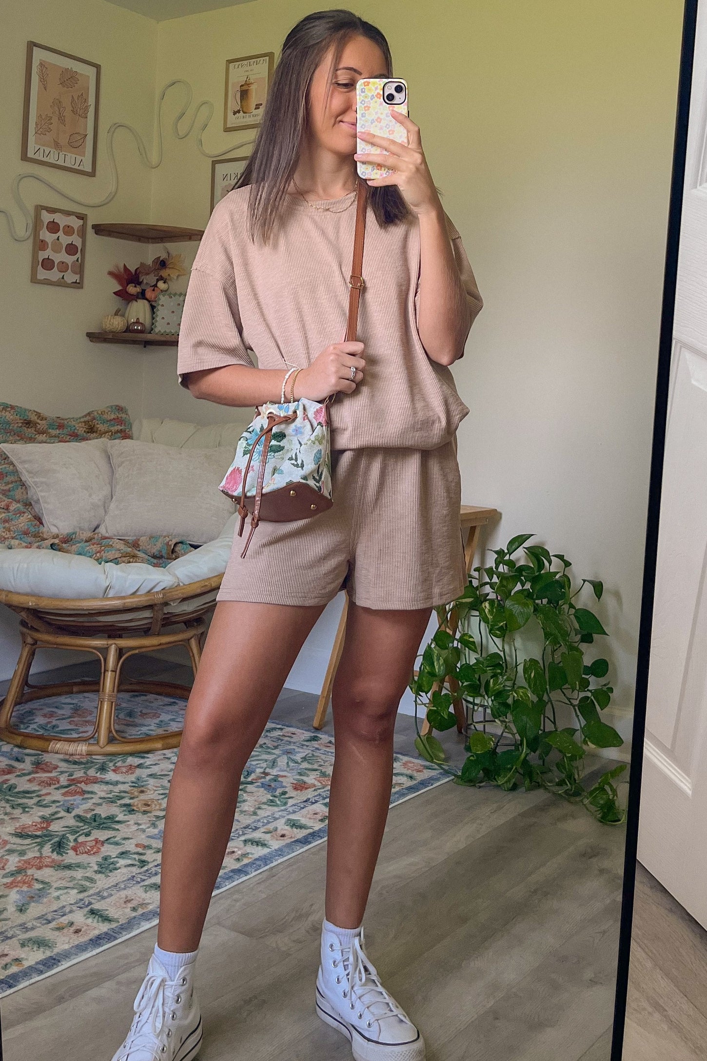 woman wearing a beige oversized short sleeve top and matching shorts, holding a floral handbag, standing in front of a mirror