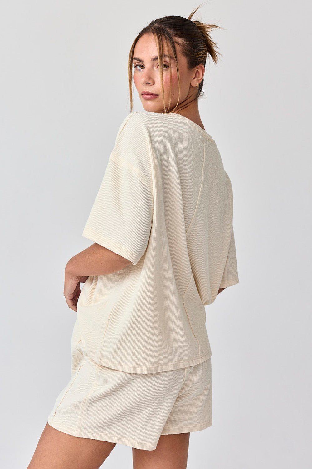 woman wearing a light beige oversized short sleeve top and matching shorts, posing with her back turned to the camera