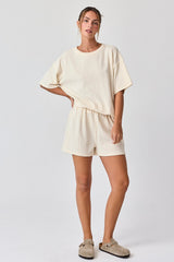 woman wearing a cream oversized short sleeve lounge set with matching shorts and casual slippers