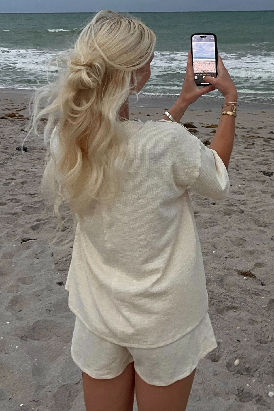 woman wearing a cream short sleeve cozy set, standing on the beach and taking a photo with her phone