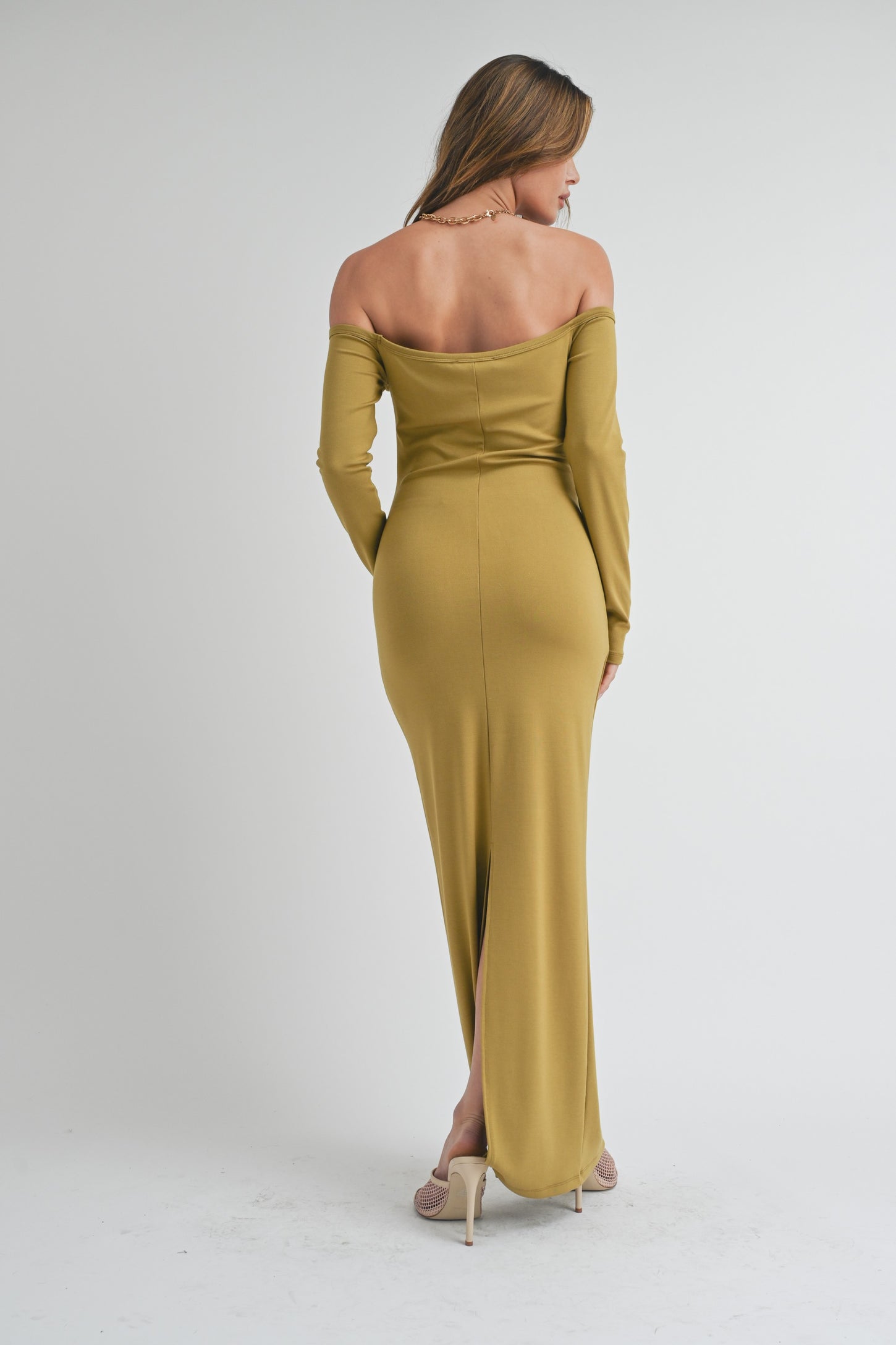 woman wearing an olive green off-shoulder maxi dress with a back slit, standing with her back to the camera