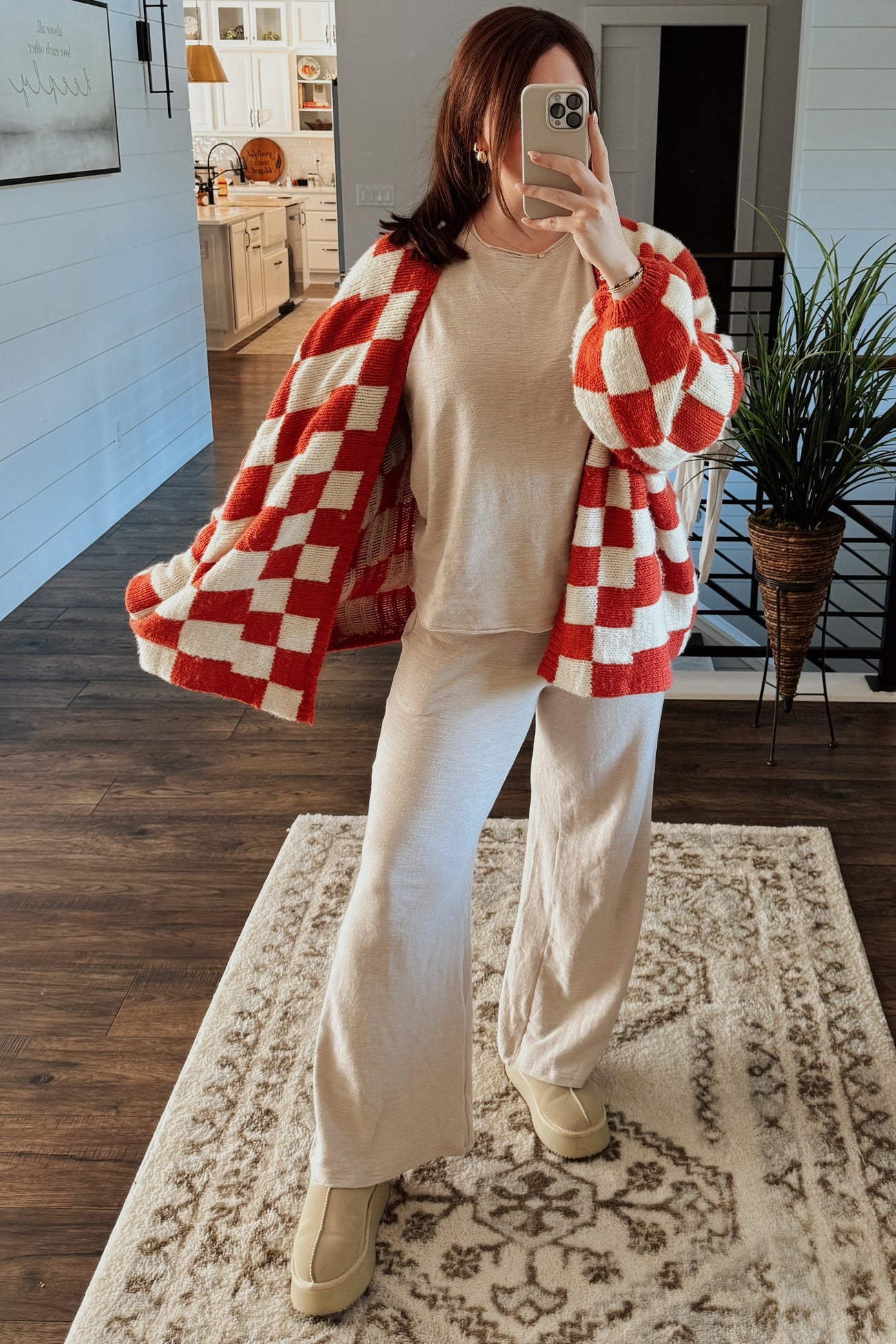 woman wearing a red and white checkered cardigan over a beige long sleeve top and matching beige pants, standing indoors