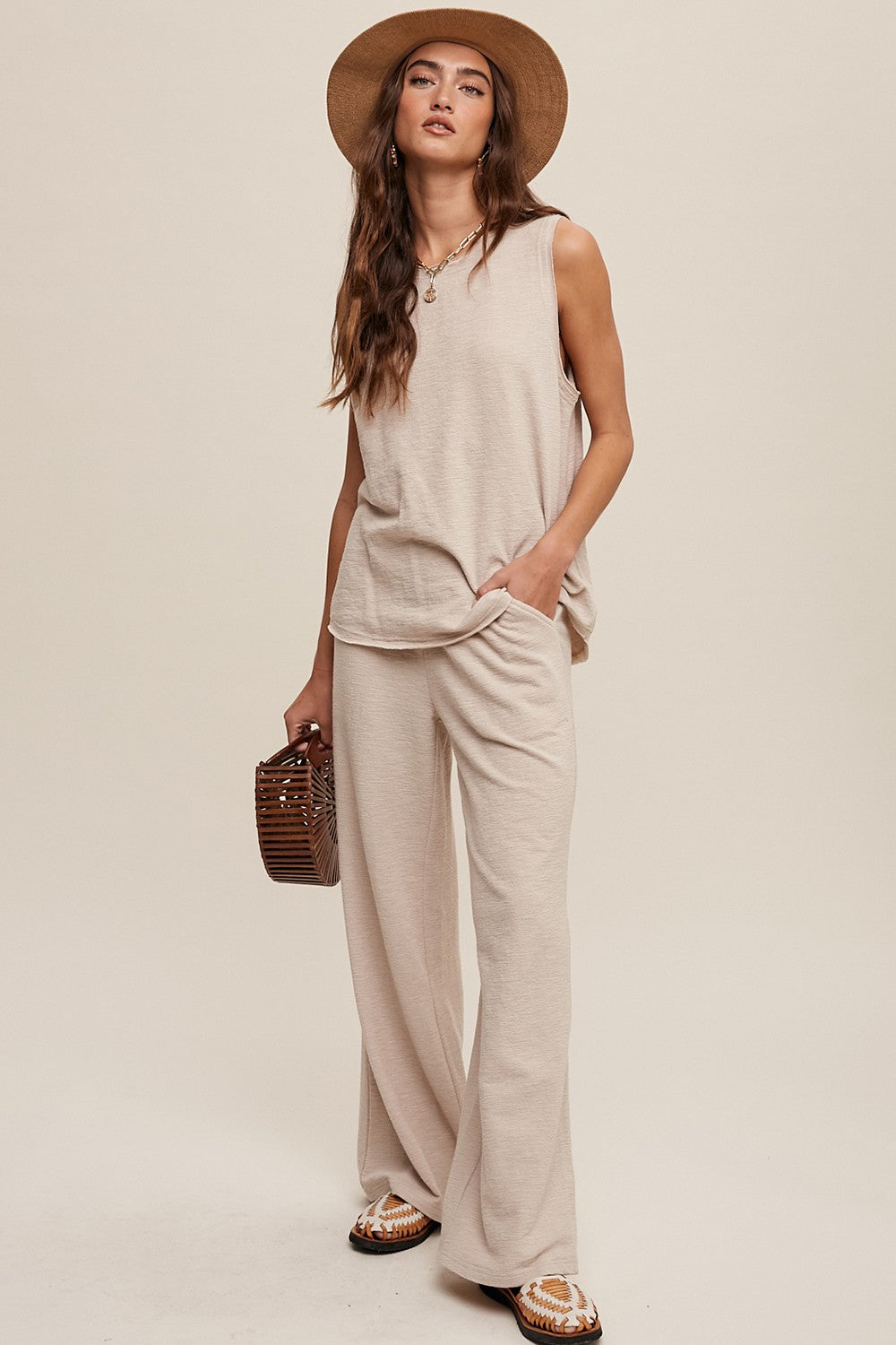 woman wearing a beige sleeveless top and wide-leg pants, accessorized with a hat and a brown handbag