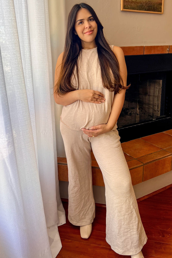 woman wearing a beige sleeveless maternity jumpsuit and holding her baby bump