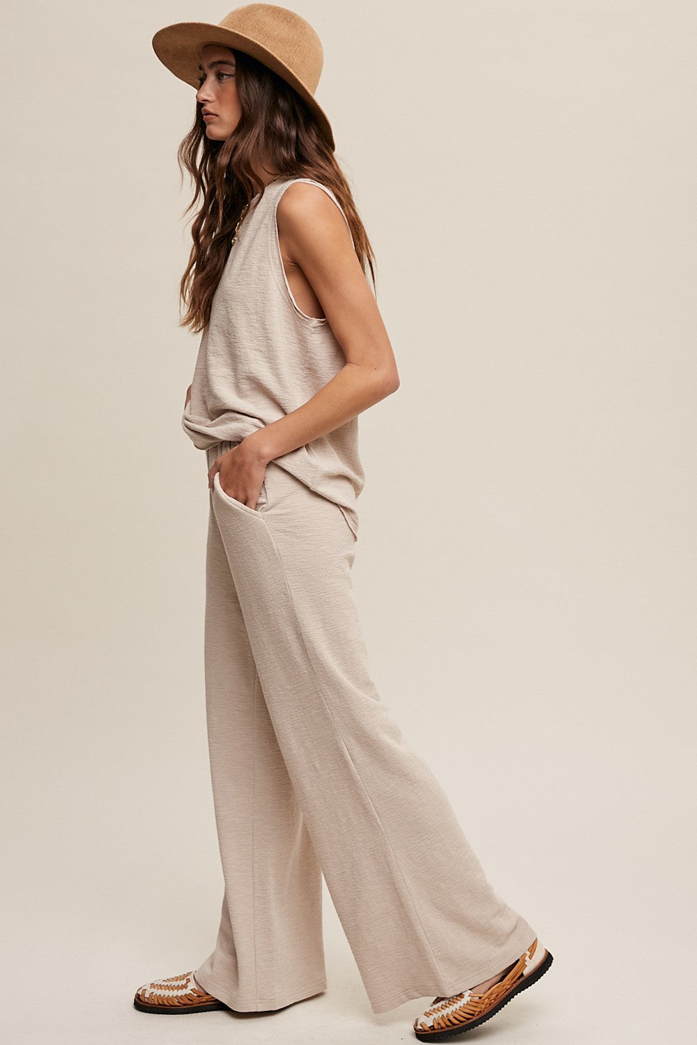 woman wearing a beige sleeveless top and matching wide-leg pants, paired with a straw hat and sandals
