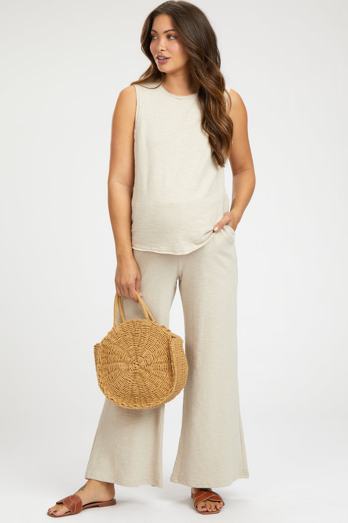 woman wearing a beige maternity sleeveless top and matching wide-leg pants, holding a woven handbag