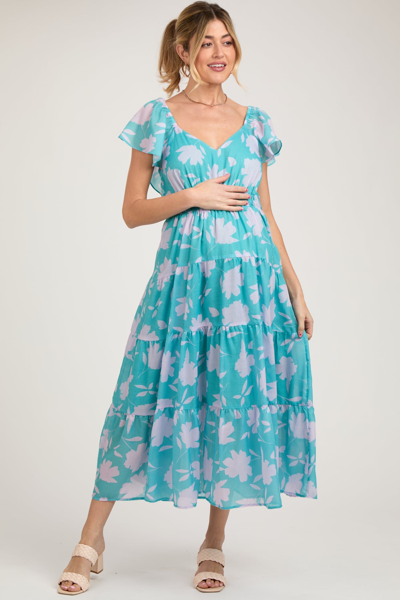 woman wearing a turquoise floral maternity dress with ruffle sleeves and a flowing tiered design, holding her baby bump