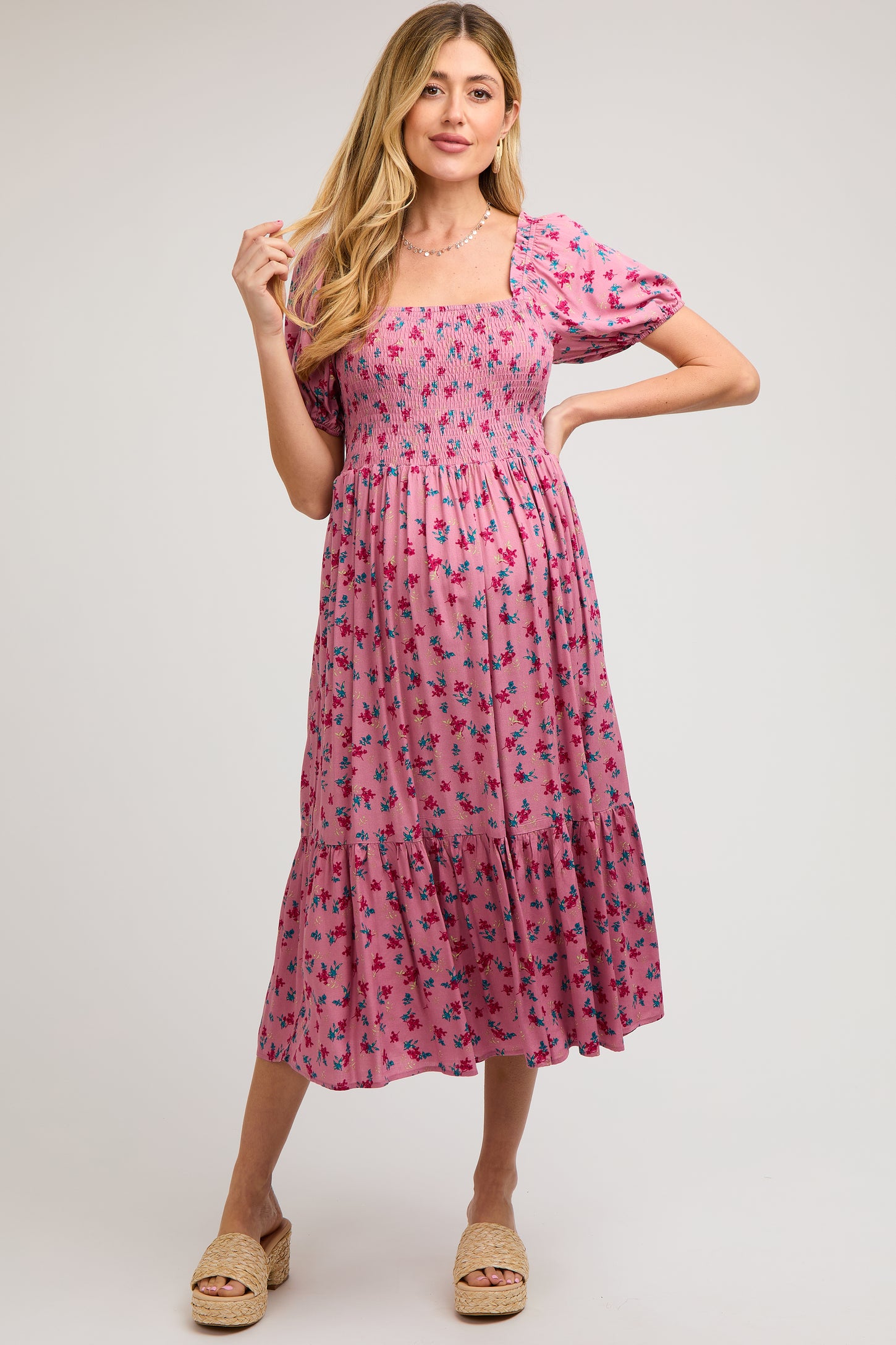 woman wearing a pink floral maternity dress with short puff sleeves and a smocked bodice, posing with a relaxed smile