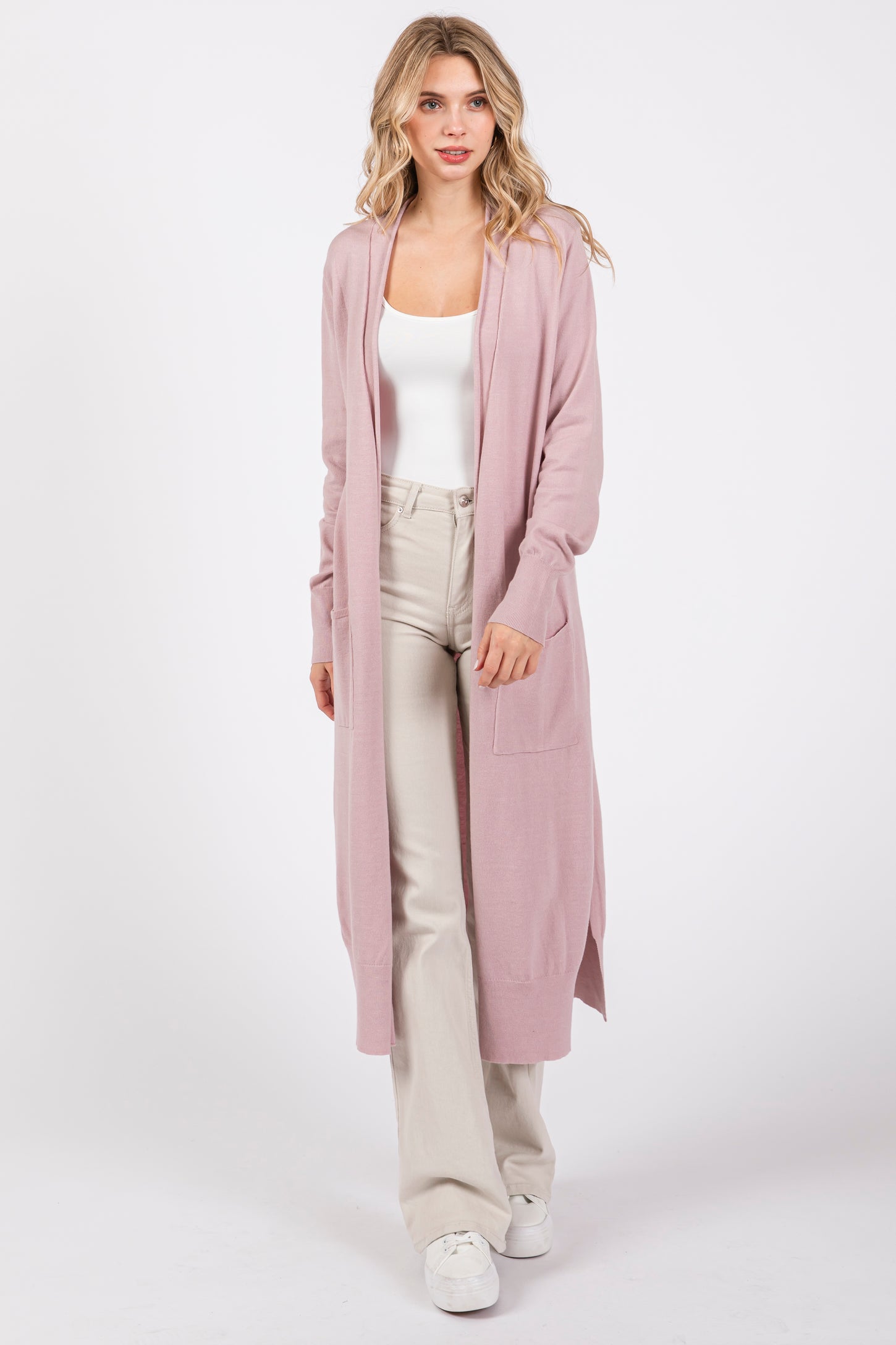 woman wearing a light pink long cardigan with pockets, paired with a white tank top and cream wide-leg pants