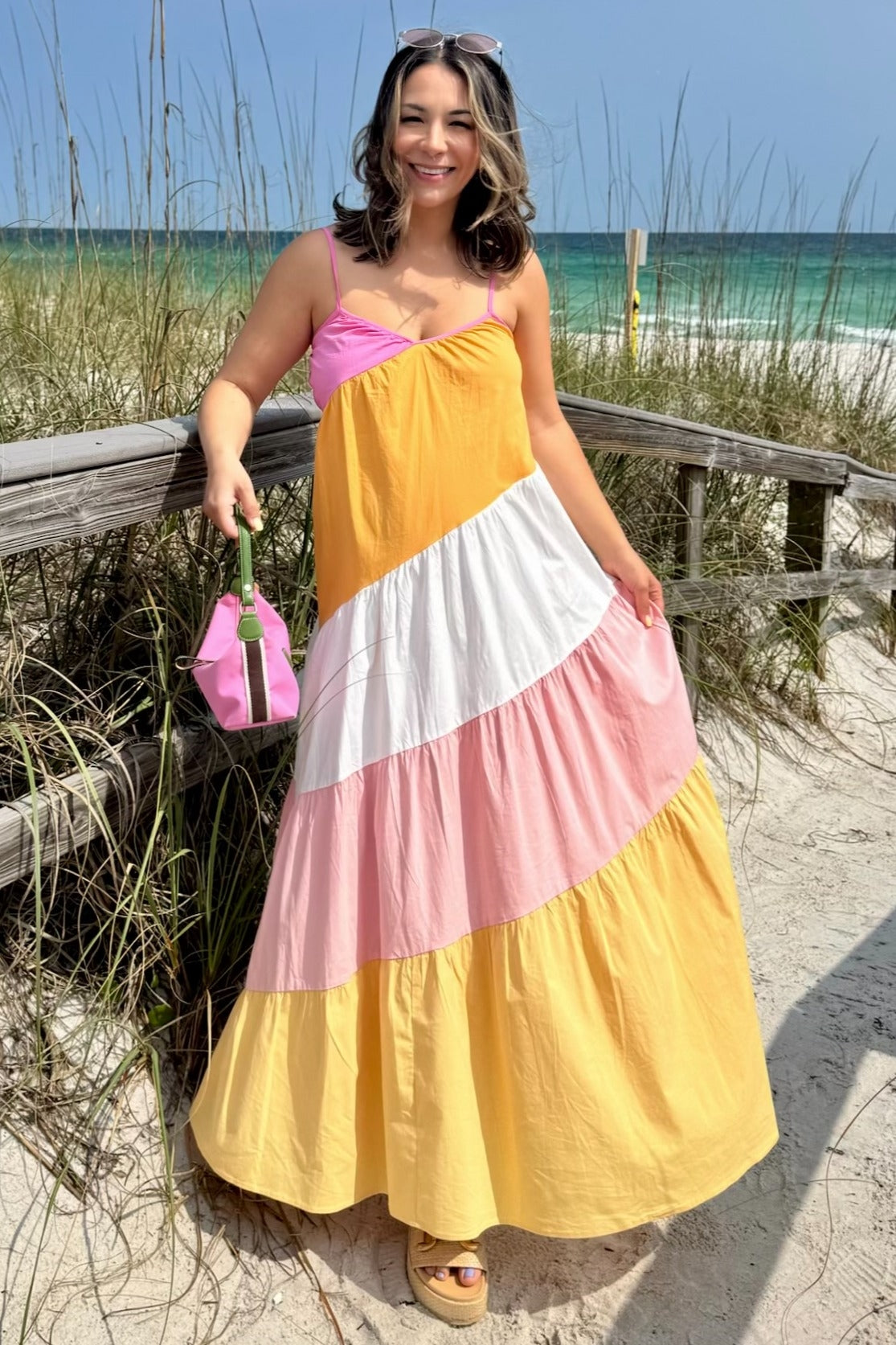 woman wearing a color-blocked maternity maxi dress with pink, white, and yellow panels, holding a mini bag and smiling at the beach