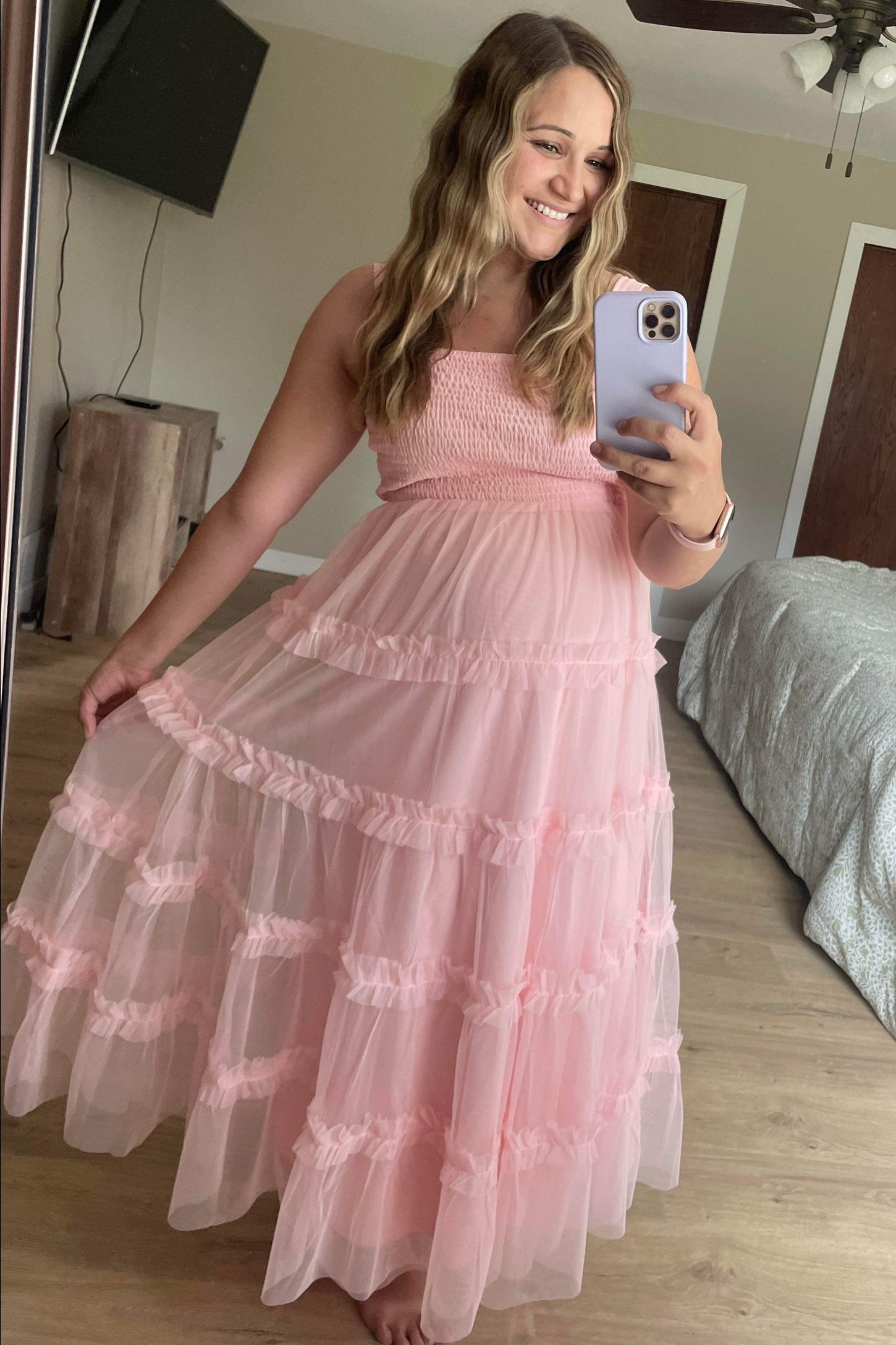 woman wearing a light pink maternity dress with ruffled layers and a fitted bodice, smiling and holding her phone in front of a mirror