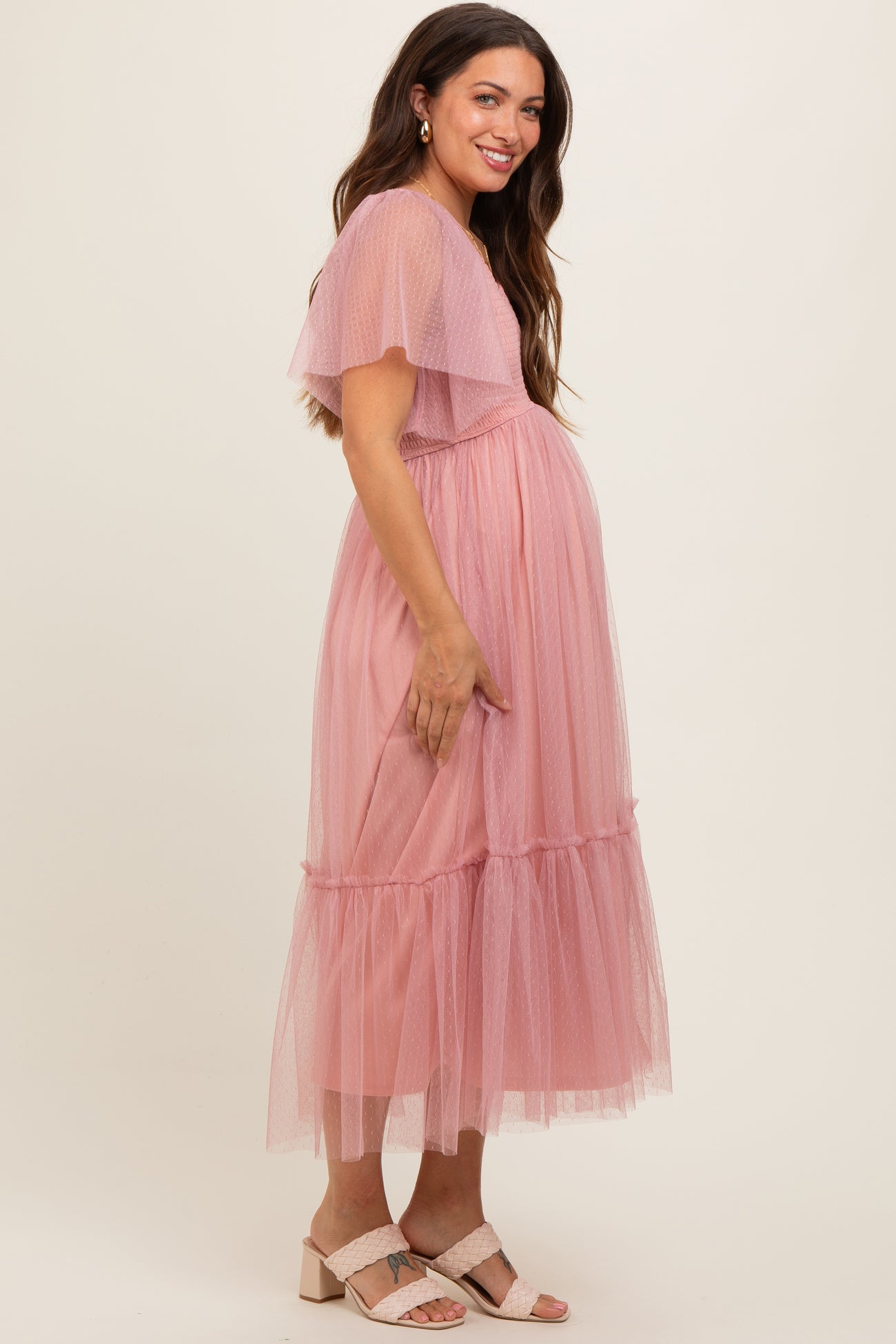 woman wearing a pink tulle maternity dress with flutter sleeves and a tiered hem, showing her baby bump