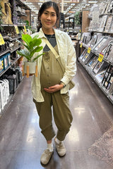 woman wearing a light green maternity jumpsuit and holding a potted plant while showing her baby bump in a store