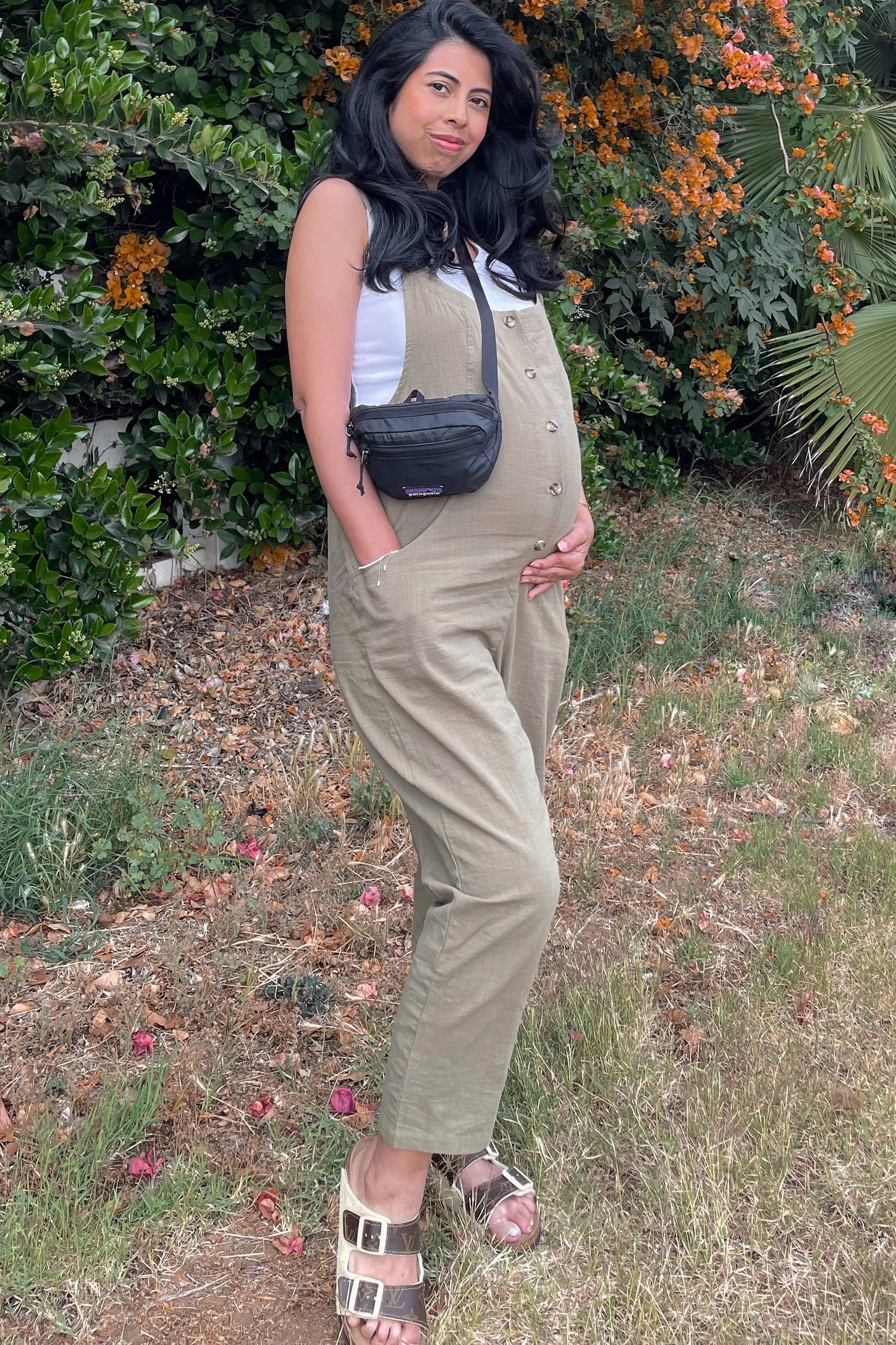 woman wearing a light olive green maternity jumpsuit and holding her baby bump in a floral outdoor setting