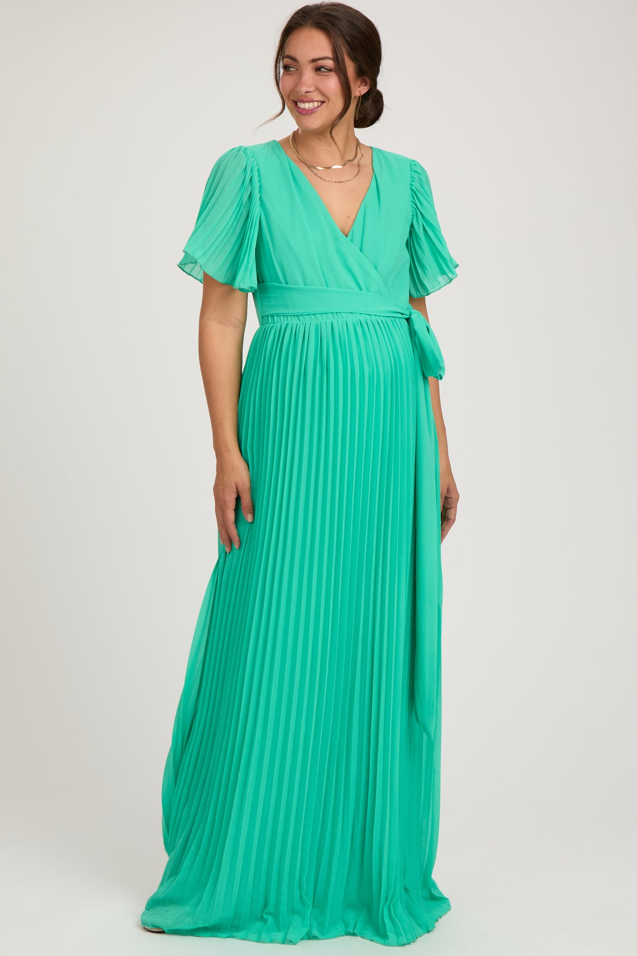 woman wearing a green pleated maternity maxi dress with short flutter sleeves and a tie waist, showing her baby bump