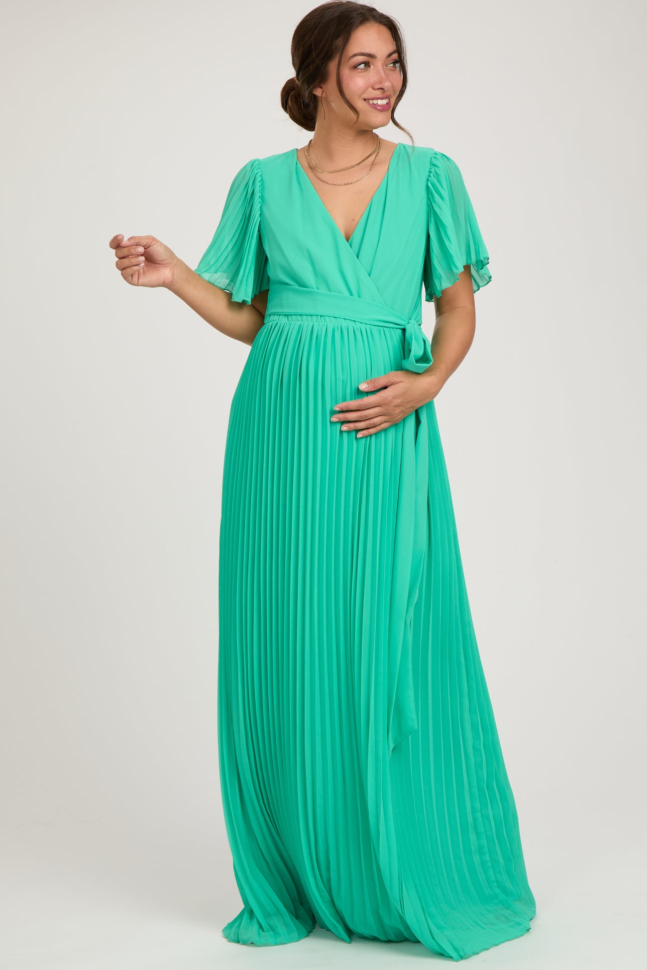 woman wearing a green pleated maternity dress with short flutter sleeves, gently cradling her baby bump