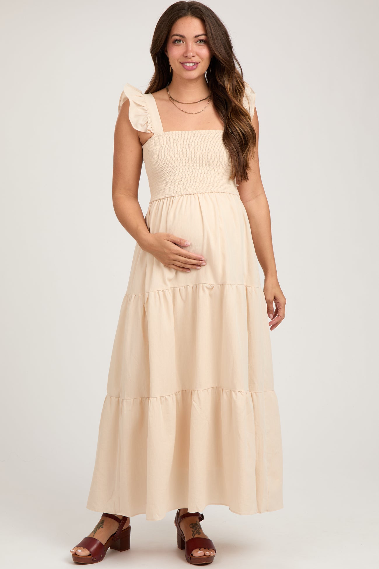 woman wearing a beige maternity maxi dress with ruffled straps and a smocked bodice, holding her baby bump