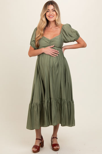 woman wearing a green maternity maxi dress with puff sleeves and a tiered skirt, holding her baby bump