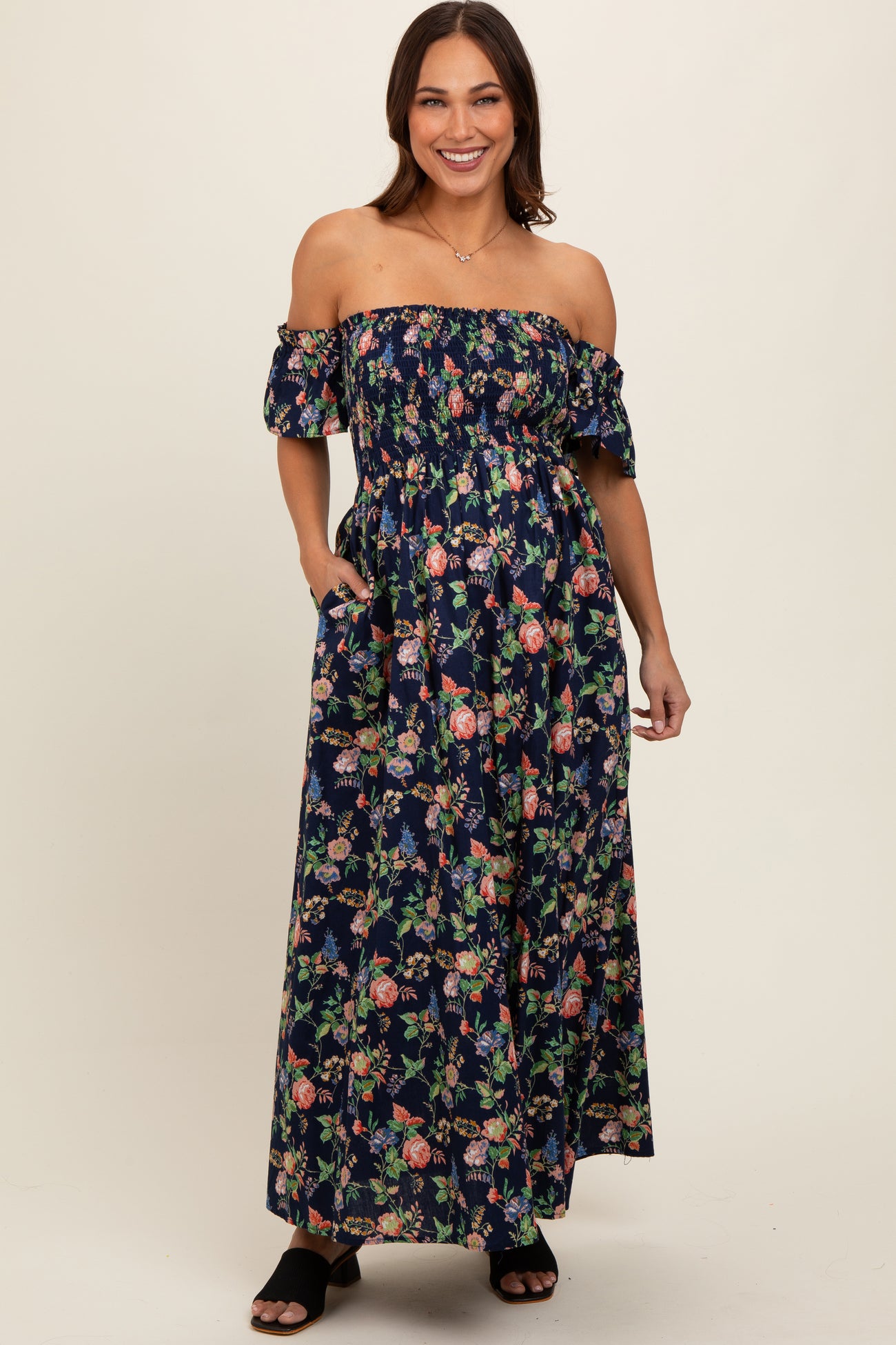 woman wearing a navy floral off-shoulder maternity maxi dress with pockets and a fitted bodice, displaying a baby bump