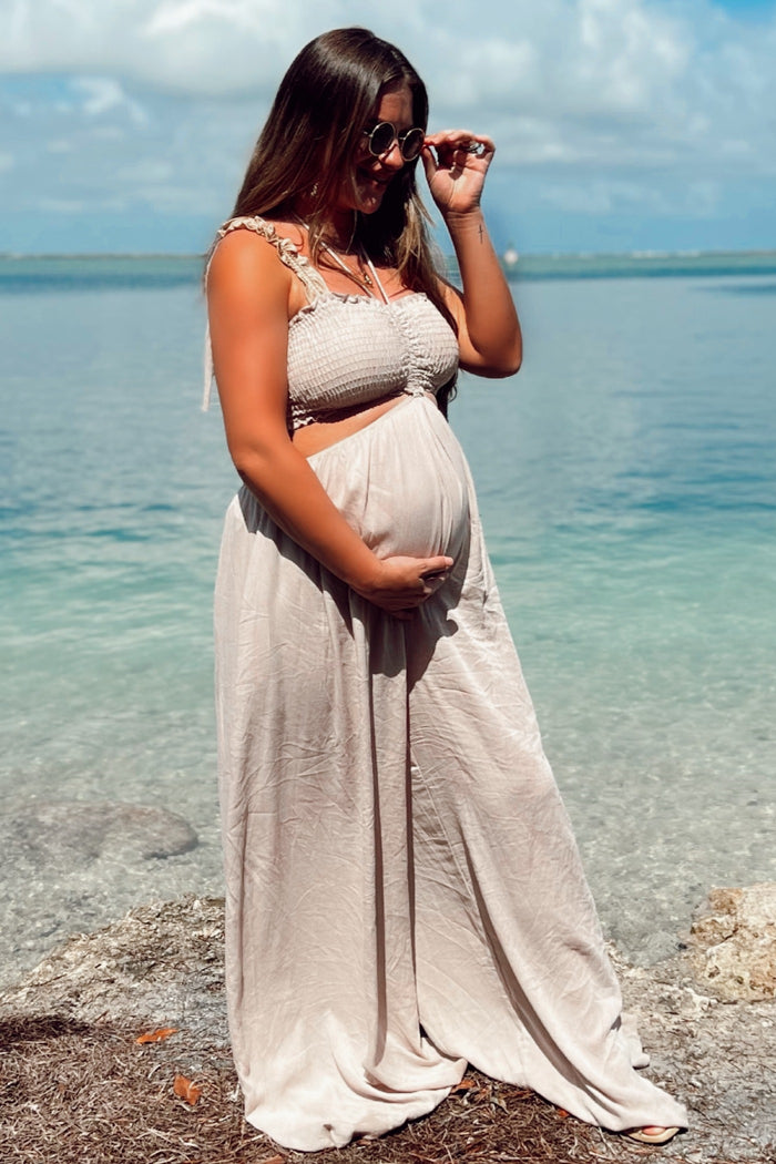 woman wearing a beige maternity maxi dress, standing by the water and gently cradling her baby bump