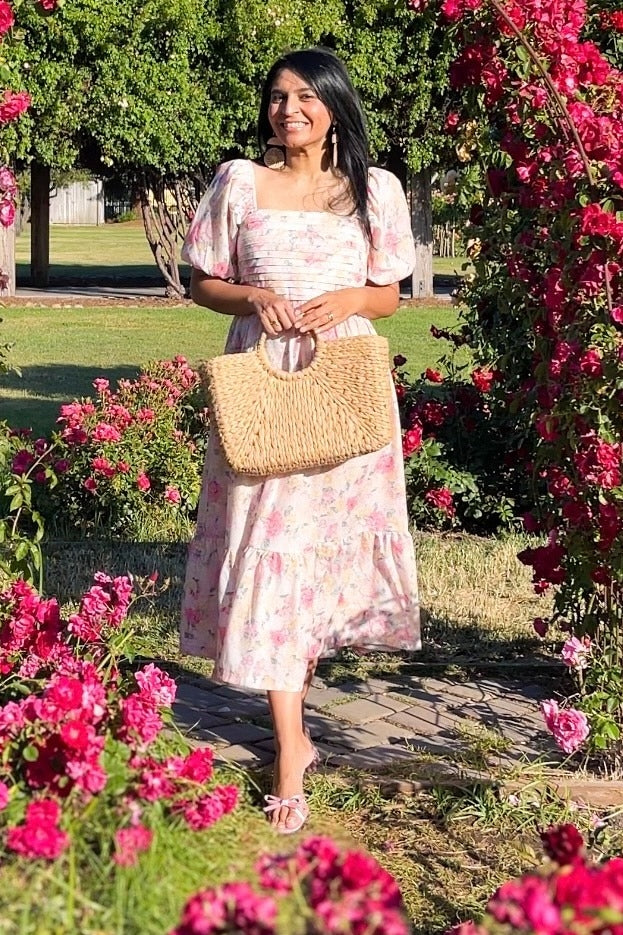 woman wearing a floral midi dress with puff sleeves and a tiered skirt, holding a woven handbag in a garden full of flowers