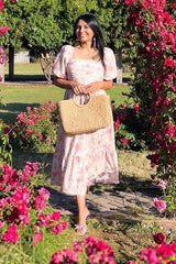 woman wearing a floral midi dress with puff sleeves and a tiered skirt, holding a woven handbag in a garden full of flowers