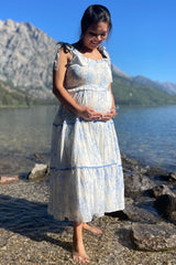 woman wearing a blue and white floral maternity dress with tiered design and holding her baby bump by the water