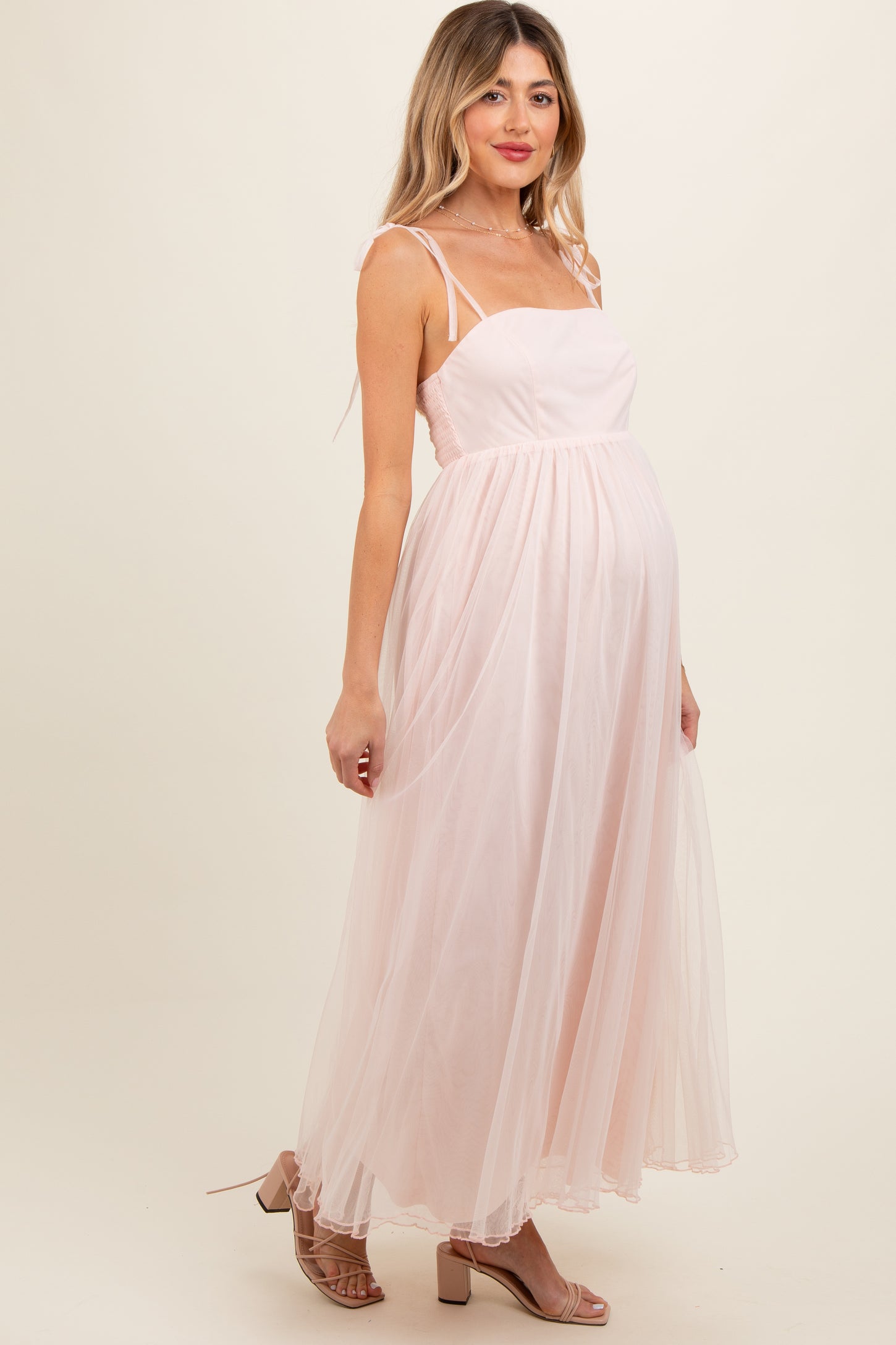 woman wearing a light pink maternity maxi dress with spaghetti straps and a flowing tulle skirt, showcasing her baby bump