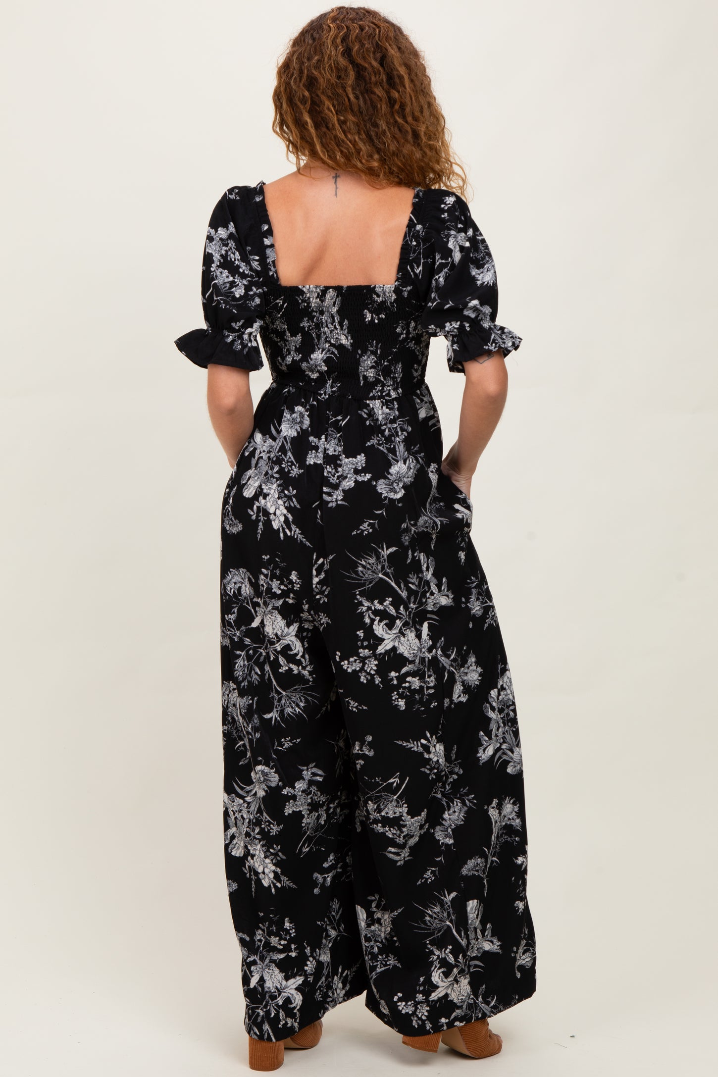 woman wearing a black floral print jumpsuit with short puff sleeves and a square back, showing the back view