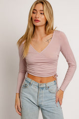 woman wearing a light pink long sleeve crop top with adjustable side ties and v-neckline, paired with high-waisted jeans