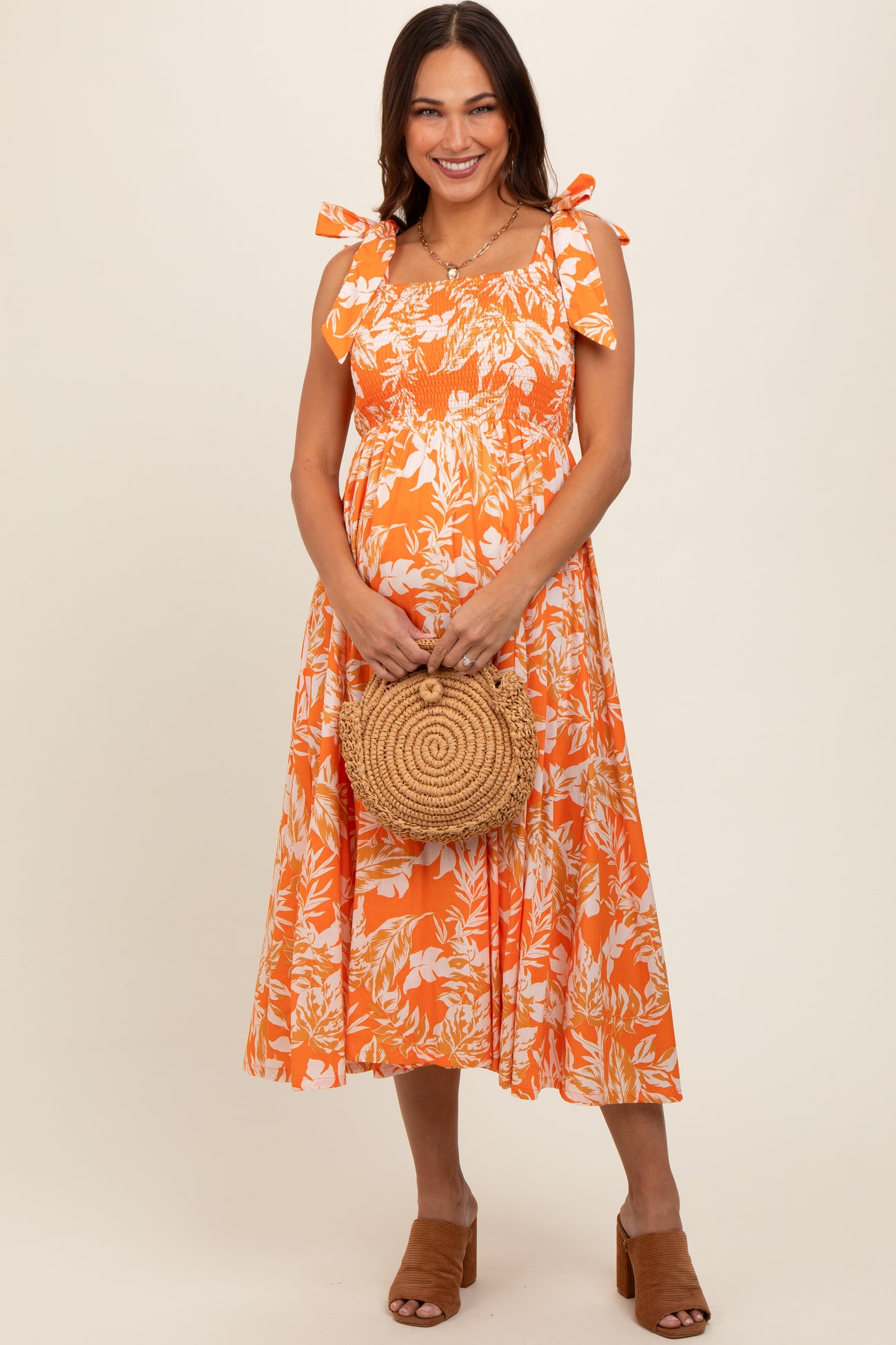 woman wearing an orange floral maternity dress with tie straps and a baby bump, holding a round straw bag
