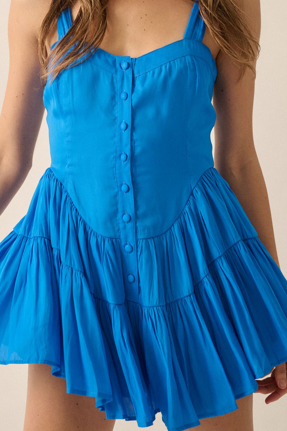 woman wearing a bright blue sleeveless mini dress with button detail and a tiered ruffled skirt