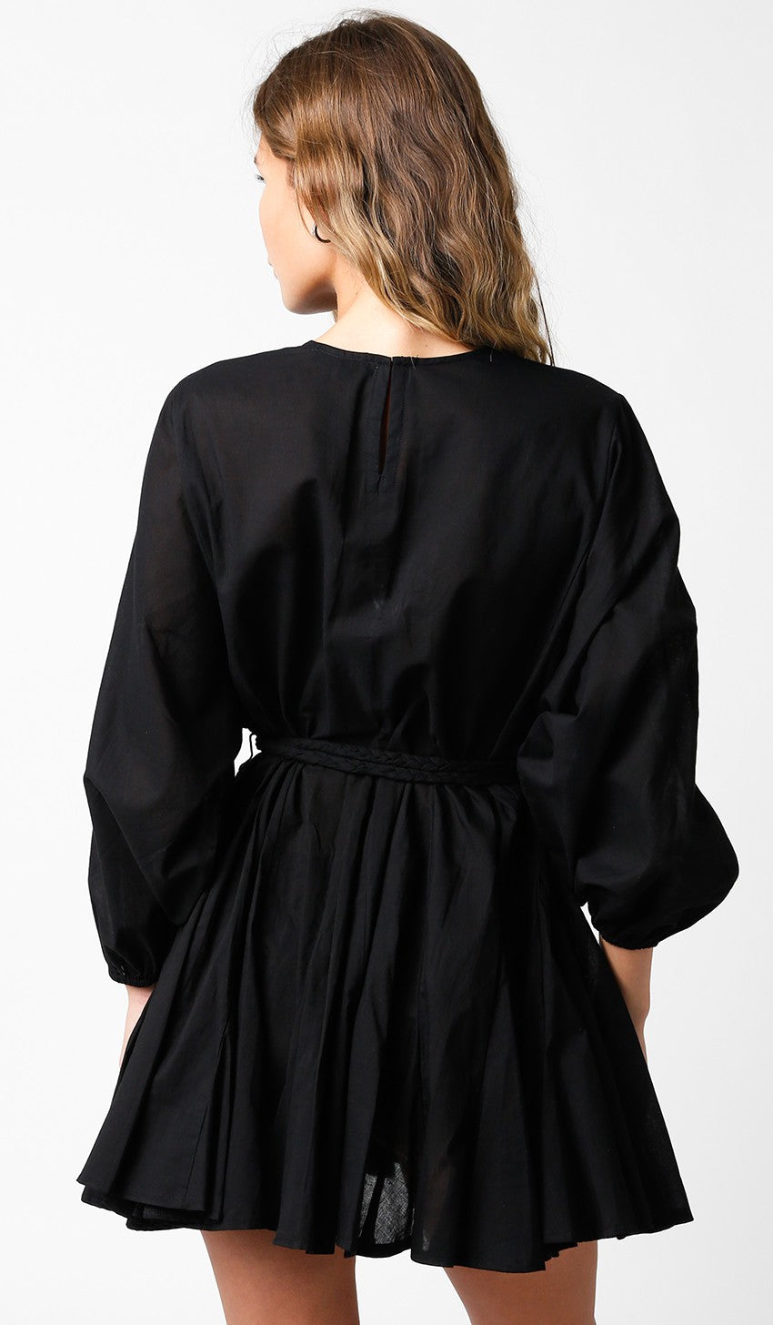 woman wearing a black belted mini dress with long sleeves and a pleated skirt, view from the back