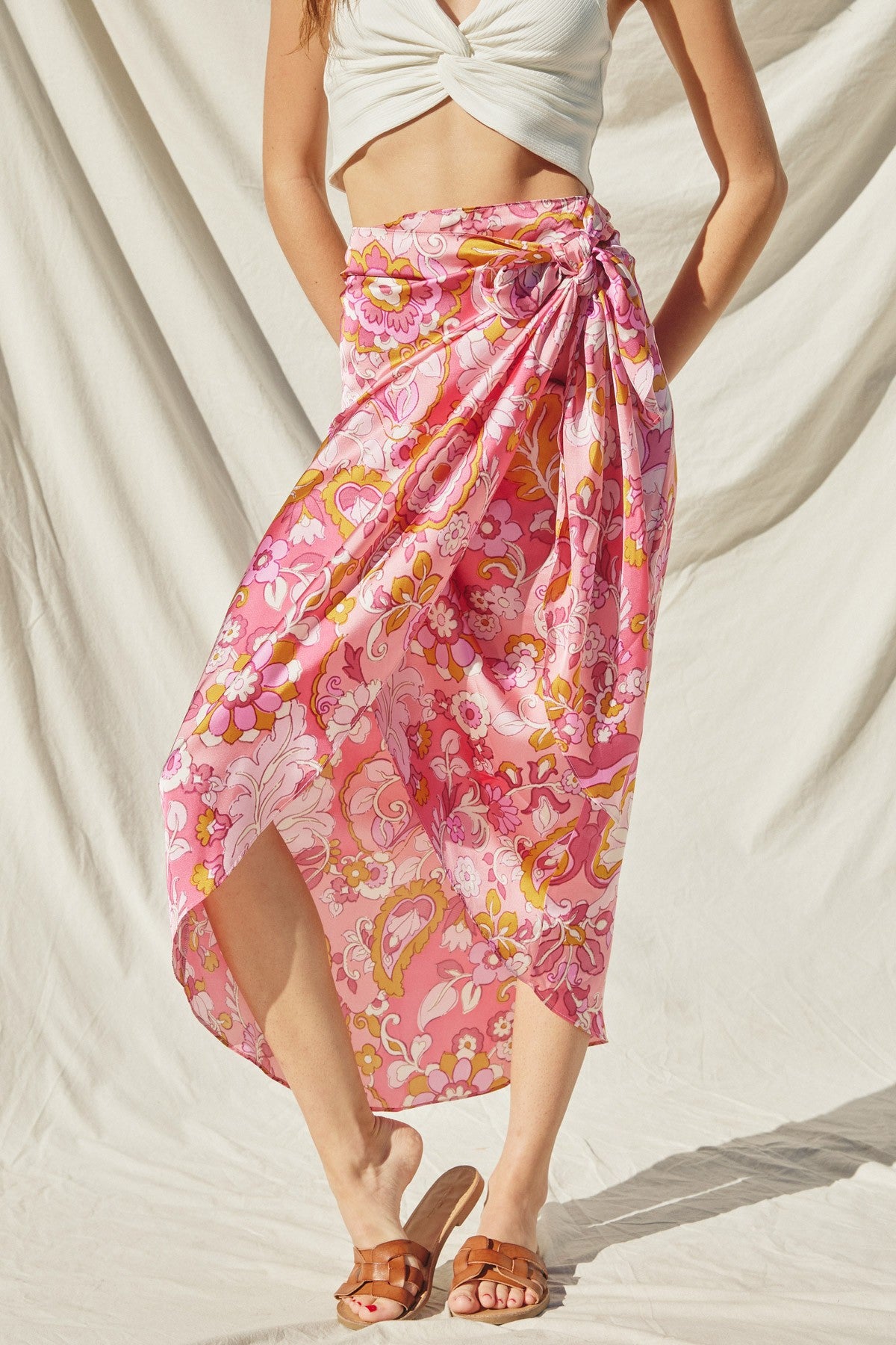 woman wearing a pink floral wrap skirt with a knotted detail, paired with a white crop top and brown sandals