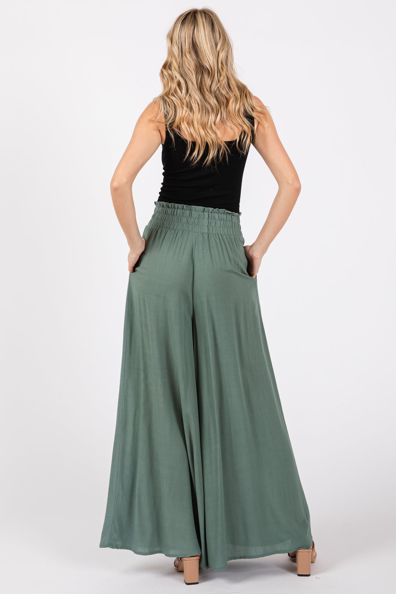 woman wearing a black tank top and green wide-leg pants, viewed from the back with hands in pockets