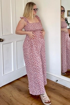 woman wearing a pink floral maternity dress with ruffle straps and a visible baby bump, standing in front of a mirror